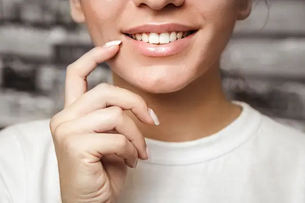 A close-up of a woman's smile as she gently touches her teeth with her finger, highlighting white and healthy teeth.